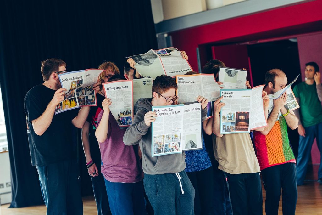 Group of 8 people standing in rows, hiding their faces whilst reading and holding up newspapers.