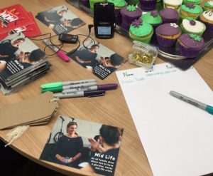 A table full of pens, swing tags, postcards and cakes, with a voice recorder ready to interview women.