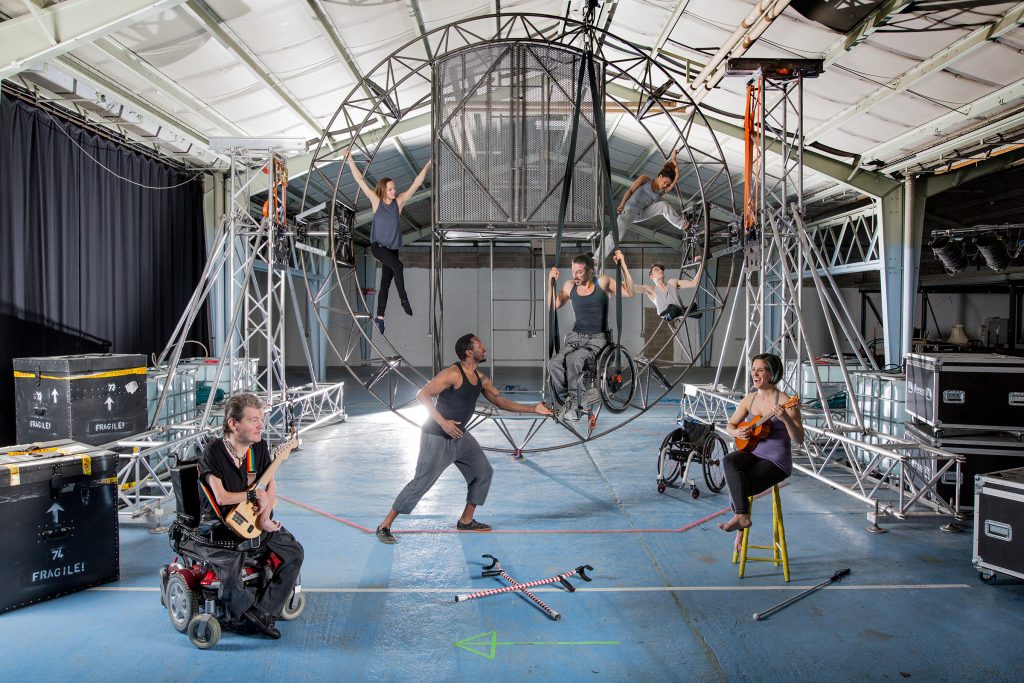 The 7 Extraordinary Bodies artists posing and moving around the new aerial set; a large circular structure suspended from both sides, with a cage-like feature and a ladder in the centre.