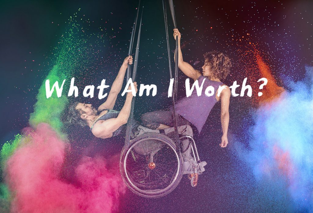 A white man in a wheelchair is suspended from aerial straps, with a strong black woman kneeling from his lap, pulling herself up on aerial straps. There is powdered paint billowing around the two circus artists in pink, green, blue and orange. The text 'What Am I Worth' is across the middle of the photograph.