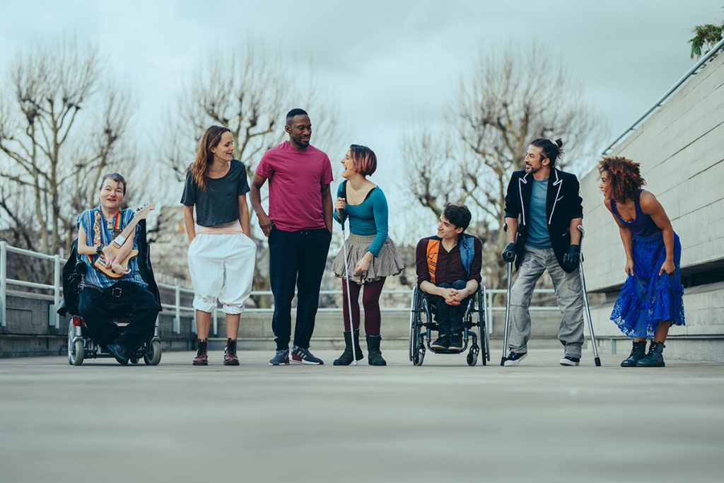 7 extraordinary artists stand in a line; left to right: a white man in a wheelchair holding a guitar, a white woman with long brown hair, a tall black man in a bright pink top, a white woman with a short bob and a walking stick, a white man in a wheelchair, a man with hair tied up holding crutches, a black woman in bold blue dress laughing.