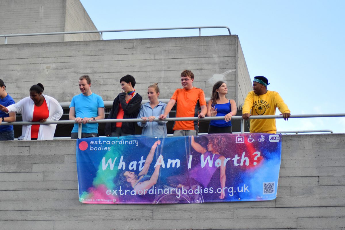 The community choir watch on and sing from the balcony above the stage - their colourful clothes are bright and empowering on the grey backdrop. They stand above the 'what am i worth' banner.
