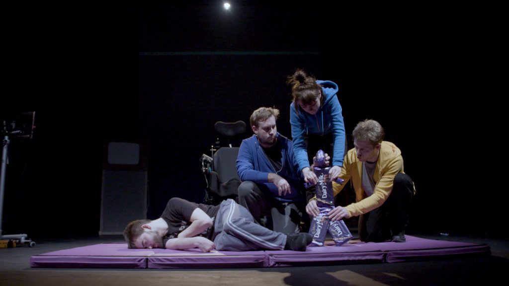 David Young lays down on a mat on the stage. Three actors are holding a 'chocolate puppet' - bars of Dairy Milk make up the body and an Easter egg makes the puppet head.